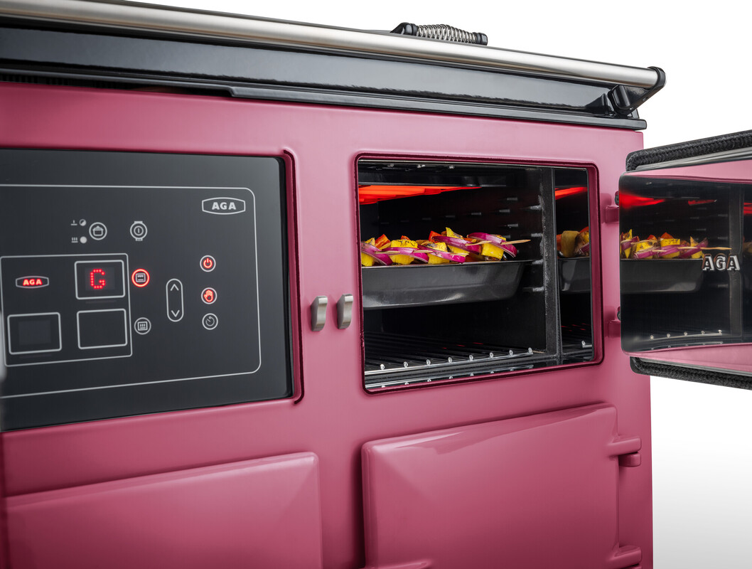 Detailaufnahme eines pinkfarbenen AGA-Ofens mit digitalen Bedienelementen und offenen Backofentüren, in denen auf Backblechen Kartoffelstücke gebacken werden