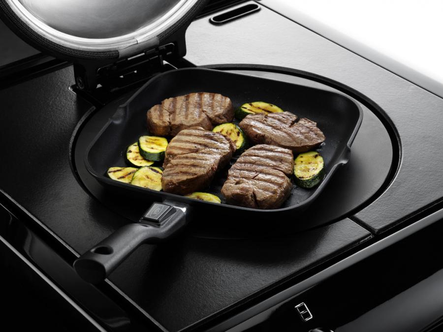 Vier gegrillte Steaks und mehrere Zucchinischeiben in einer Grillpfanne auf einem schwarzen modernen Herd