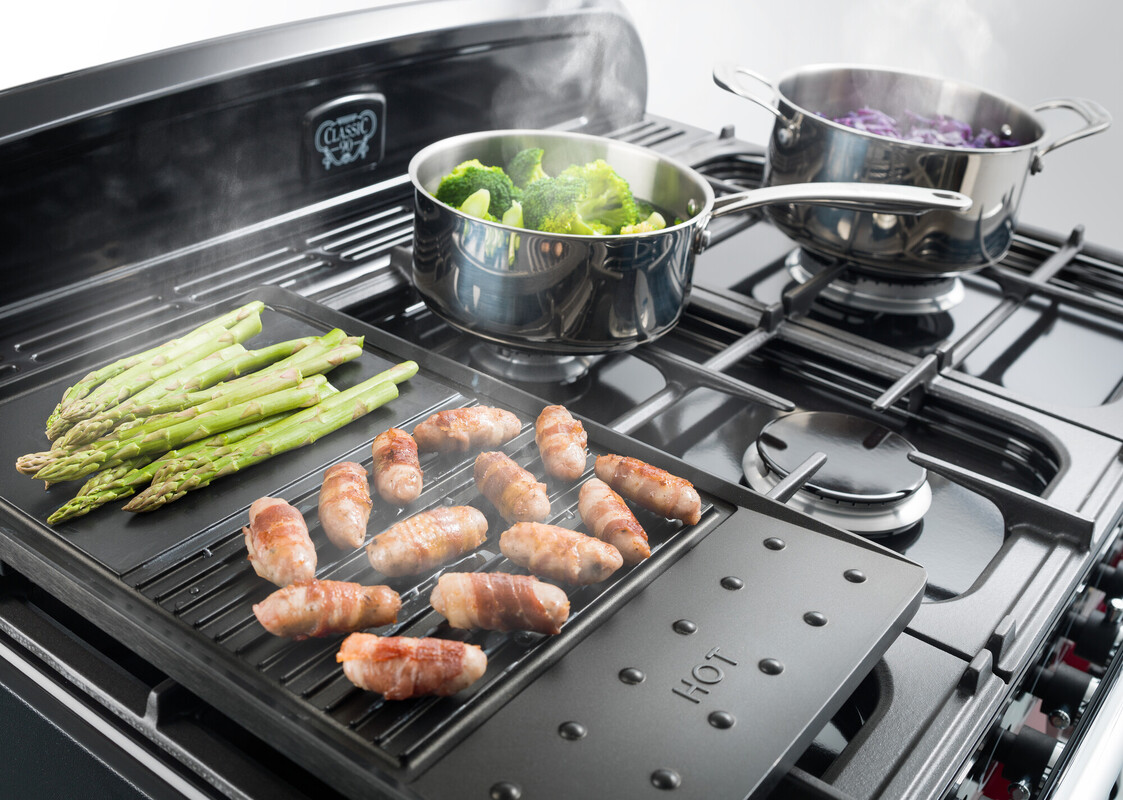 Frischer grüner Spargel und Würstchen auf einer Grillplatte auf einem Gasherd, im Hintergrund Töpfe mit dampfendem Brokkoli und Rotkohl