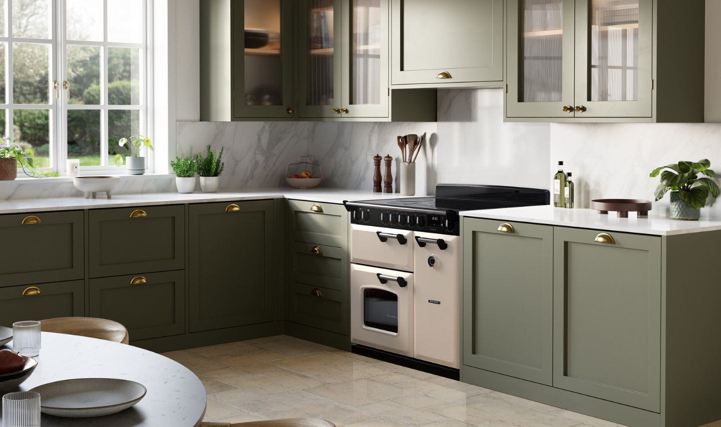 Helle Landhausküche: salbeigrüne Fronten, Glasoberschränke, weiße Marmor-Rückwand und Arbeitsplatte, cremefarbener Range Cooker, Kräuter am Fenster, Ölflaschen und Accessoires auf der Arbeitsfläche